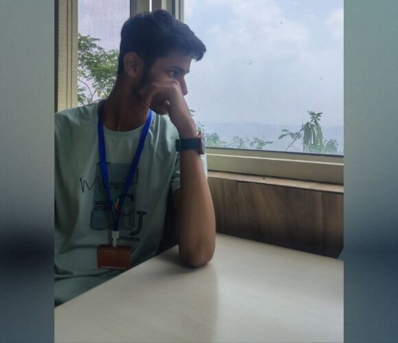 A person sitting at a table by a window, wearing a light-colored t-shirt and blue lanyard with an ID card, looking outside at a cloudy sky and greenery.